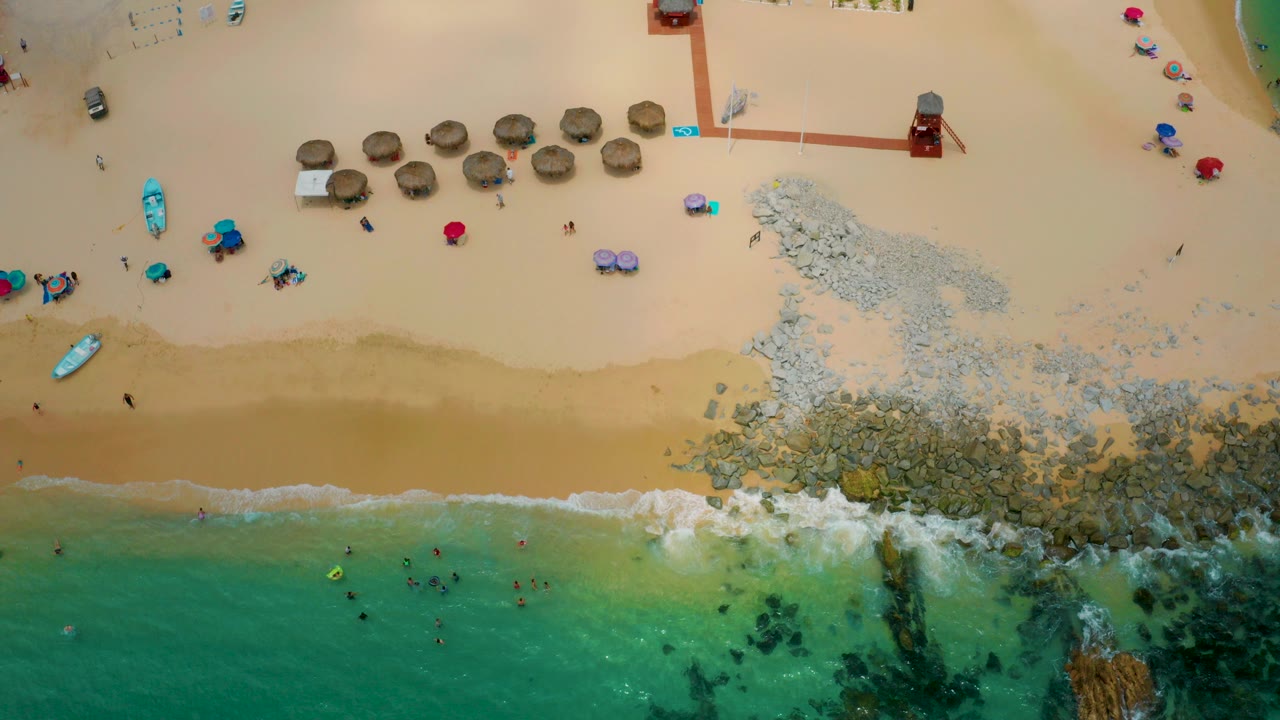 Overhead view of Palmilla Beach Cabo, Mexico with tan sand, green-blue water, a rocky shoreline patch, umbrellas, beach craft, swimmers, and scattered people on the sand.