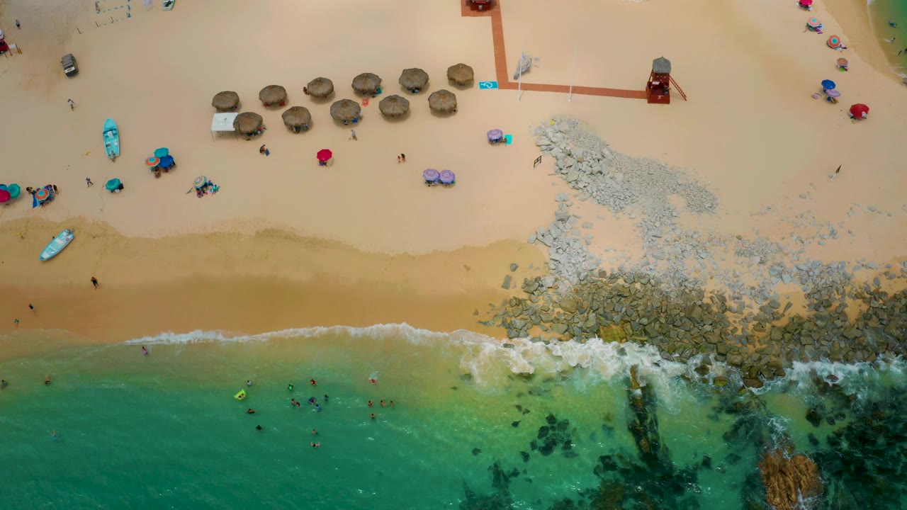 Aerial view over Palmilla Beach Cabo with pale sand across the upper frame, clear water below, swimmers offshore, foam reaching rocks, and small groups near umbrellas and beach craft.