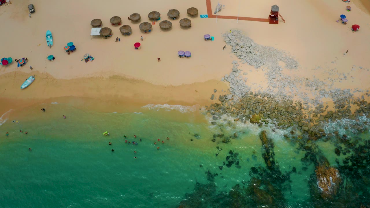 Top-down view of Palmilla Beach Cabo, Mexico with turquoise water below, tan sand above, a pale rock spread at the shoreline, umbrellas and small boats on the beach, and tiny distant people visible.