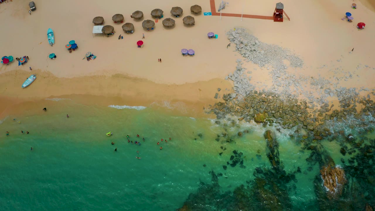 Palmilla Beach Cabo seen from above with shallow turquoise water, darker submerged rocks, a pale stone formation on the sand, rows of thatched umbrellas, and tiny beachgoers visible near the waterline.