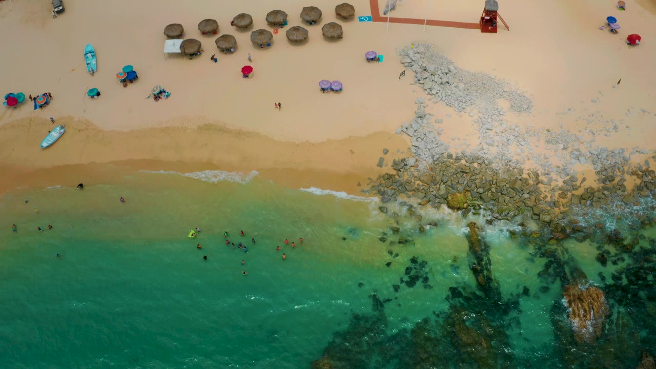 Overhead view of Palmilla Beach Cabo with waves brushing the tan beach, clustered swimmers in the lower water, rocky surf at right, and umbrellas, boats, and beach gear set back on the sand.