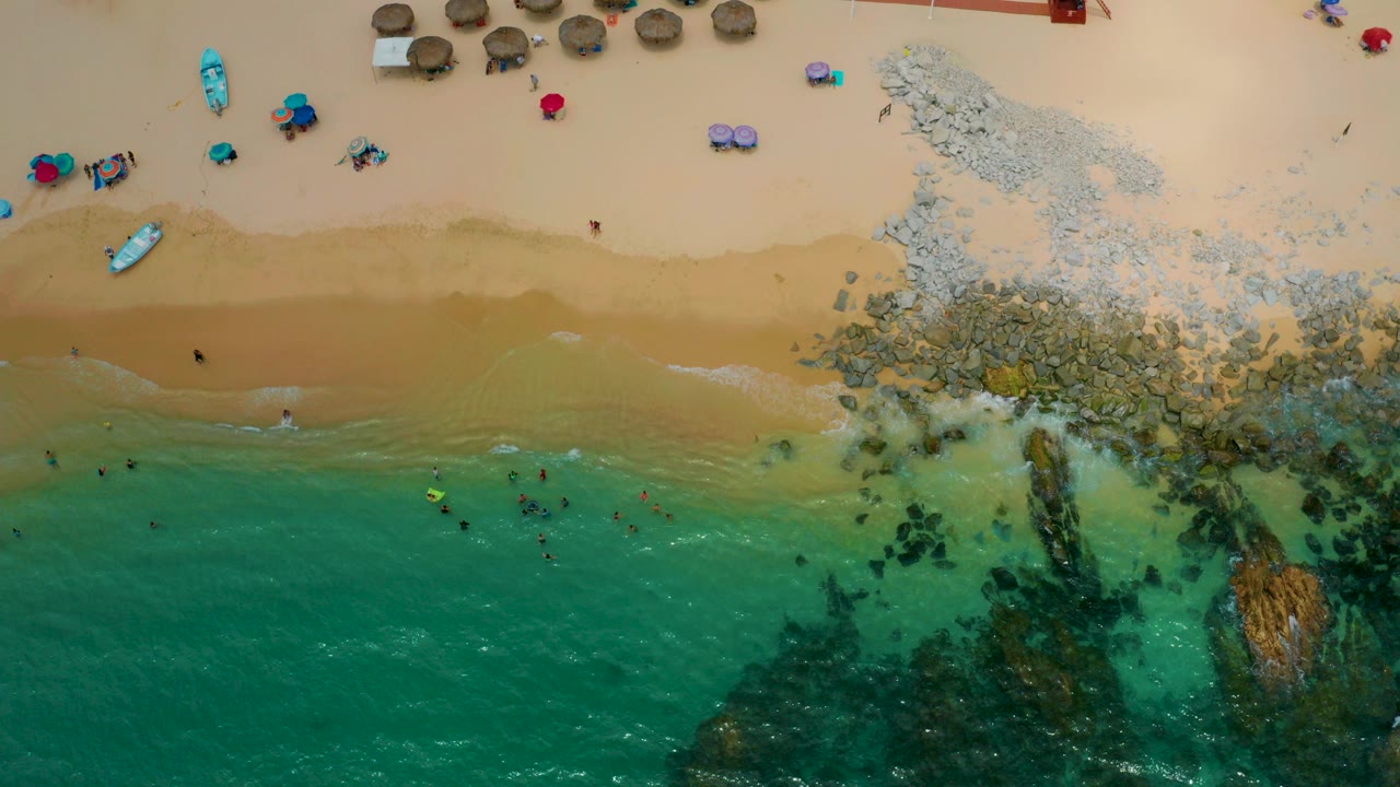 Overhead view of Palmilla Beach Cabo, Mexico, showing turquoise water, dark rocks, tan sand, swimmers near shore, umbrellas, beach chairs, and palapas at the top of the frame.
