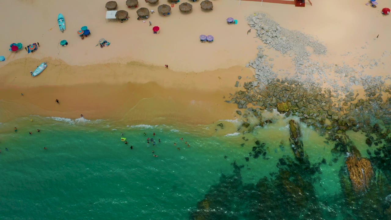 Aerial view of Palmilla Beach Cabo with a wider band of tan sand, a pale surf line, swimmers near the lower water edge, and a cluster of palapas and umbrellas near the upper beach.
