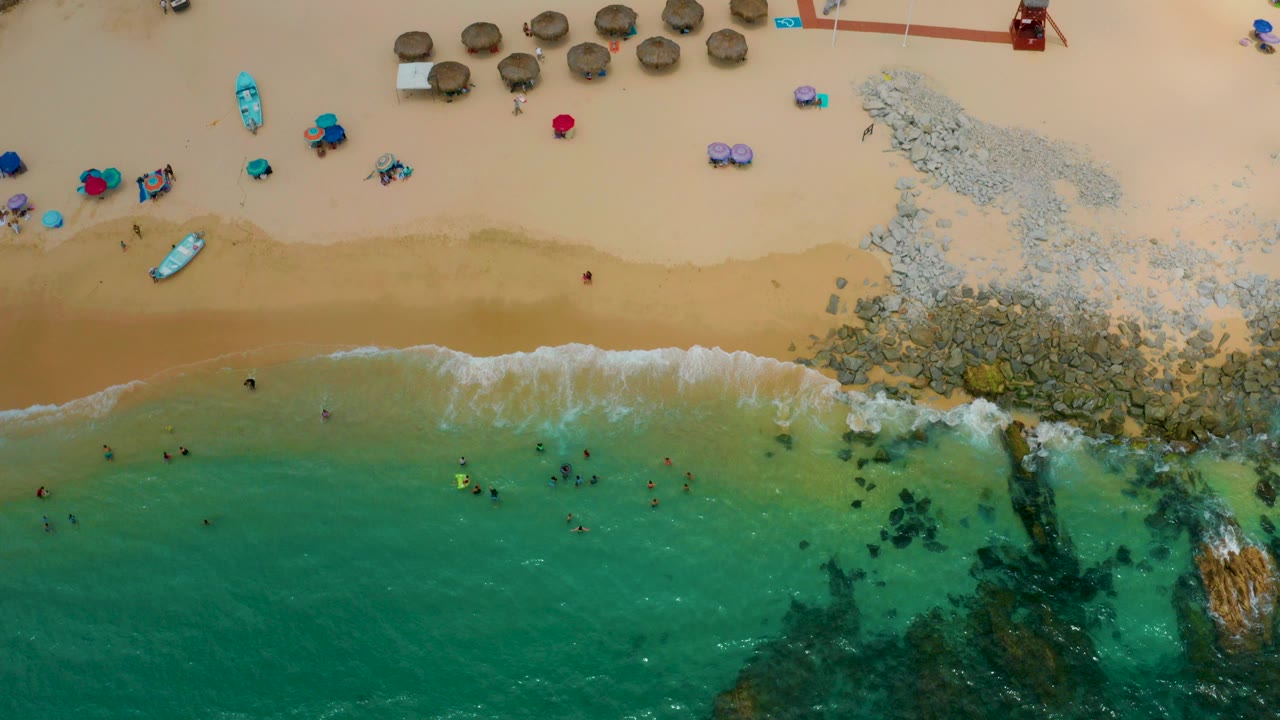 Overhead view of Palmilla Beach Cabo, Mexico showing turquoise water below, tan sand above, swimmers near the shore, thatched umbrellas, colorful beach gear, and a rocky patch at right.