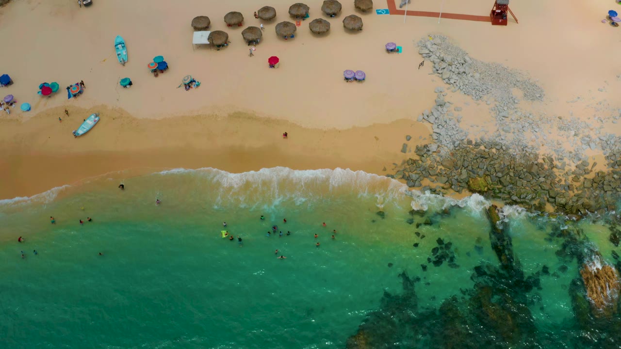 Aerial shoreline with beach umbrellas, swimmers, rocks, and shallow green water.