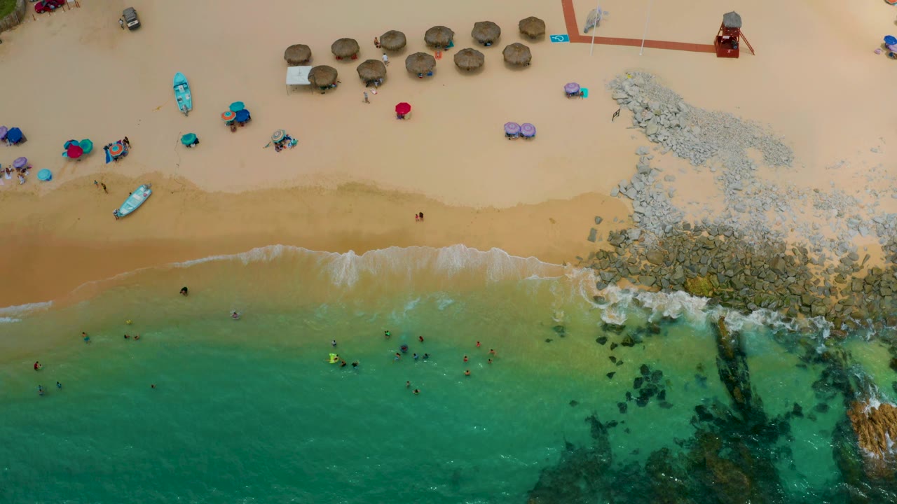 Top-down view of Palmilla Beach Cabo, Mexico with the surf line across the middle, swimmers below it, umbrellas and towels above it, rocks at right, and a small red tower near the upper edge.