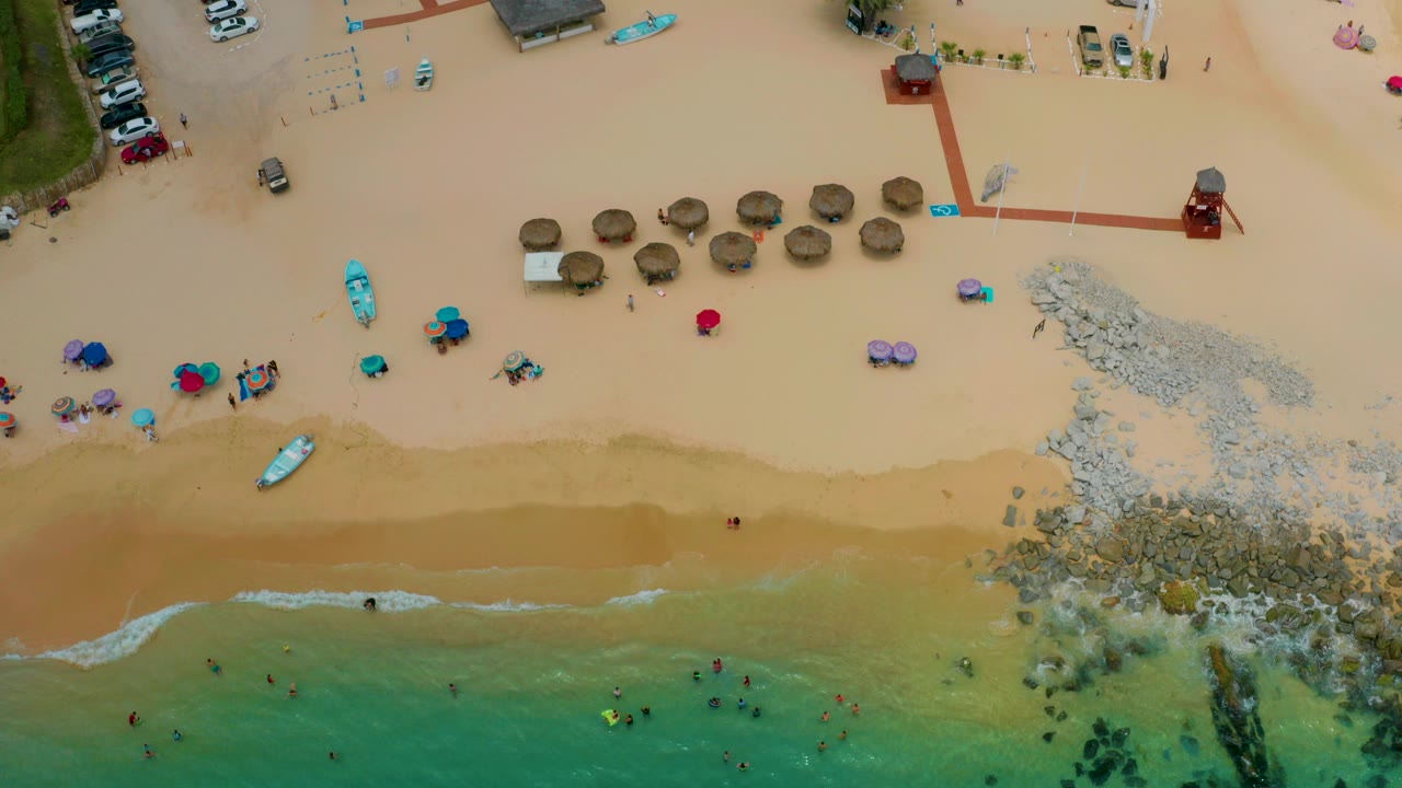 Top-down aerial frame of Palmilla Beach Cabo with green-blue water at the bottom, tan sand, palapa umbrellas, rocks, small boats, parked cars, and tiny beachgoers visible.