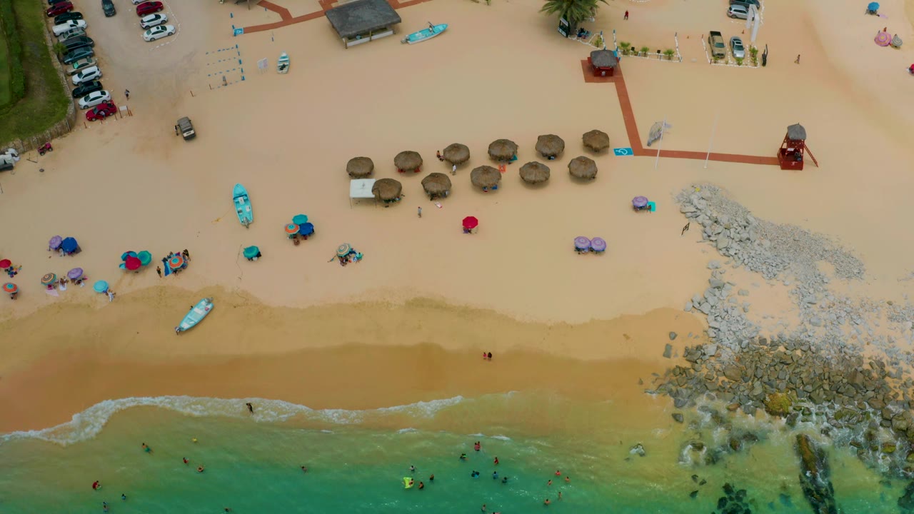 A top-down view of Palmilla Beach Cabo shows more of the beach setup, including round palapas, a red path, a rocky shoreline break, turquoise shallows, small boats, and tiny people scattered near the water.