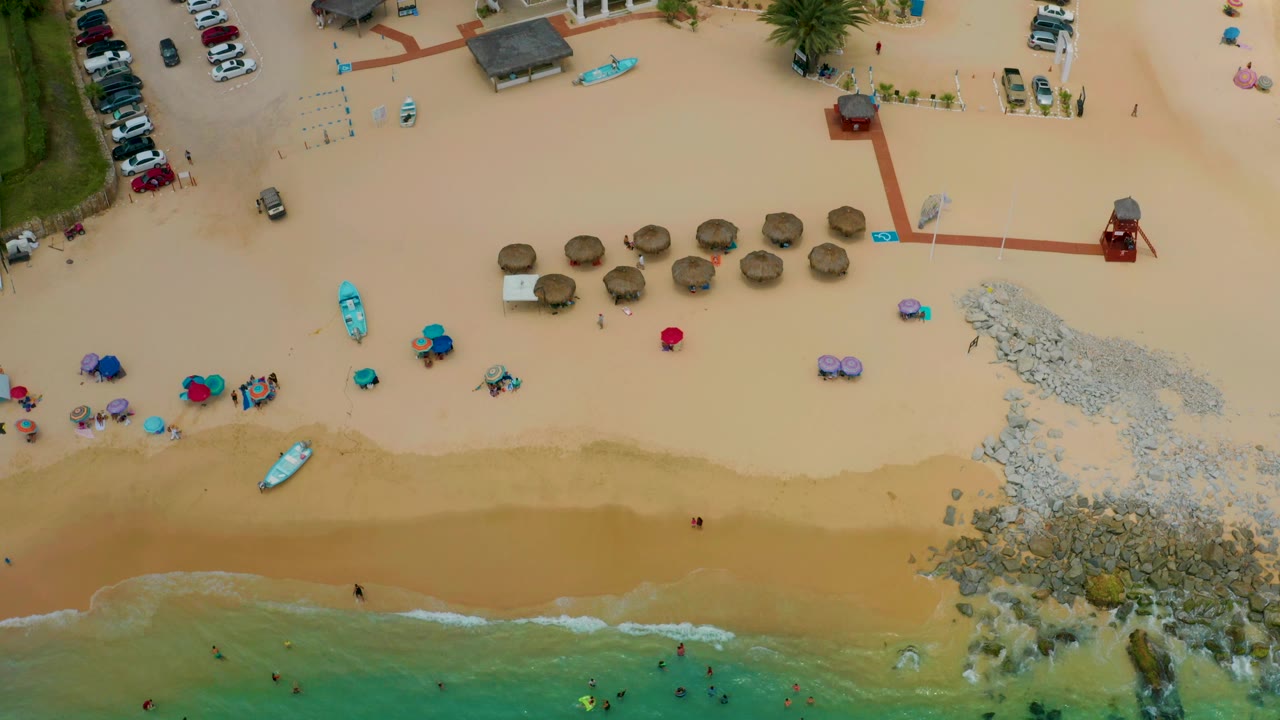 Drone view of beach surf, umbrellas, small boats, rocks, and resort-side parking.