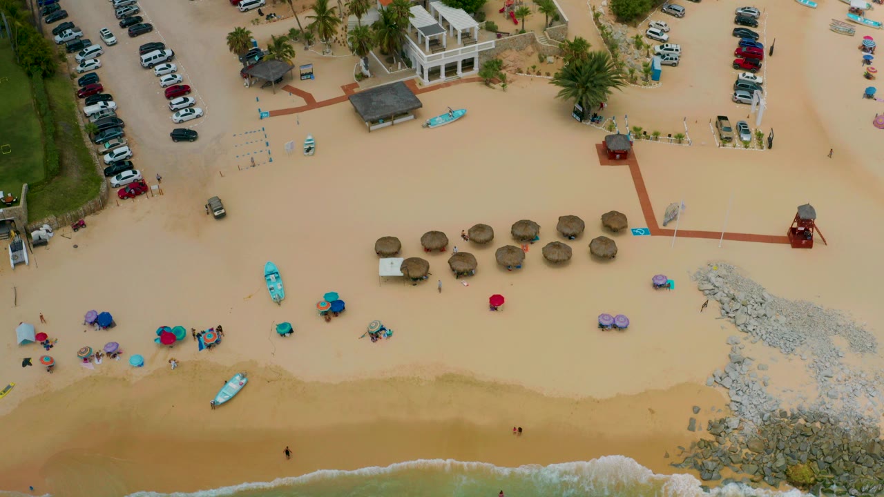 Overhead frame from Palmilla Beach Cabo, Mexico showing the shoreline at the bottom, straw shade umbrellas across the sand, turquoise kayaks, parked cars, palms, low beach buildings, and no visible people.