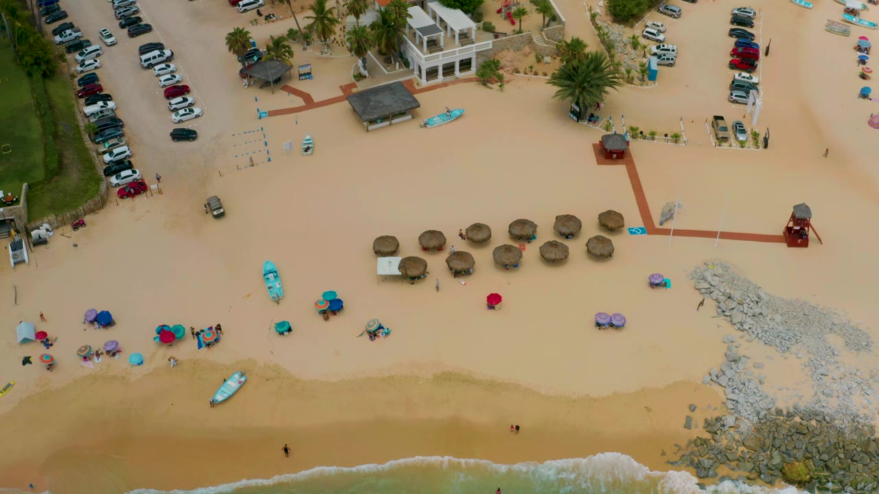 Aerial view over Palmilla Beach Cabo, Mexico with small waves along the lower sand, a row of straw umbrellas, bright kayaks, a parking area, palms, and no visible people.