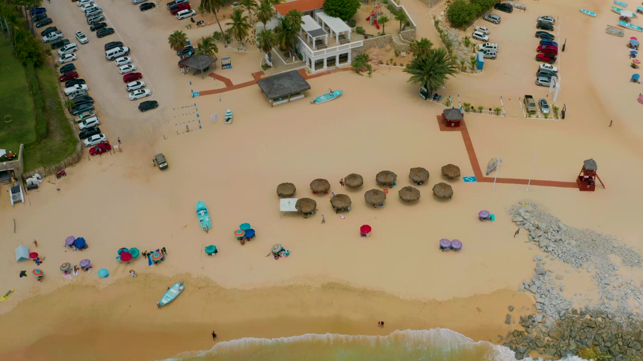 Top-down frame from Palmilla Beach Cabo, Mexico showing the surf line lower in frame, shade umbrellas centered on the sand, kayaks nearby, parking and low buildings above, and no visible people.