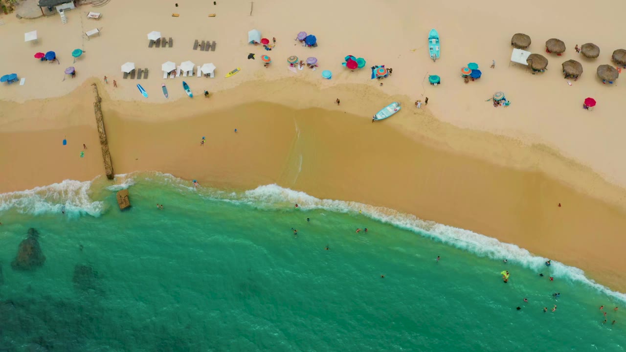 Aerial view of Palmilla Beach Cabo, Mexico at 0:00, with turquoise surf below, tan sand above, beach chairs, umbrellas, small craft, and distant people visible as tiny figures.