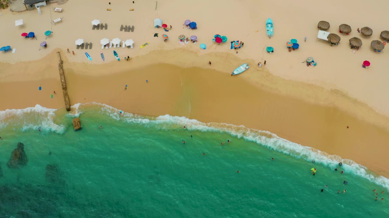 Palmilla Beach Cabo, Mexico appears from above at 0:02, with a diagonal white wave line, clear green-blue water, a beach setup on the sand, and small distant people near the shore.