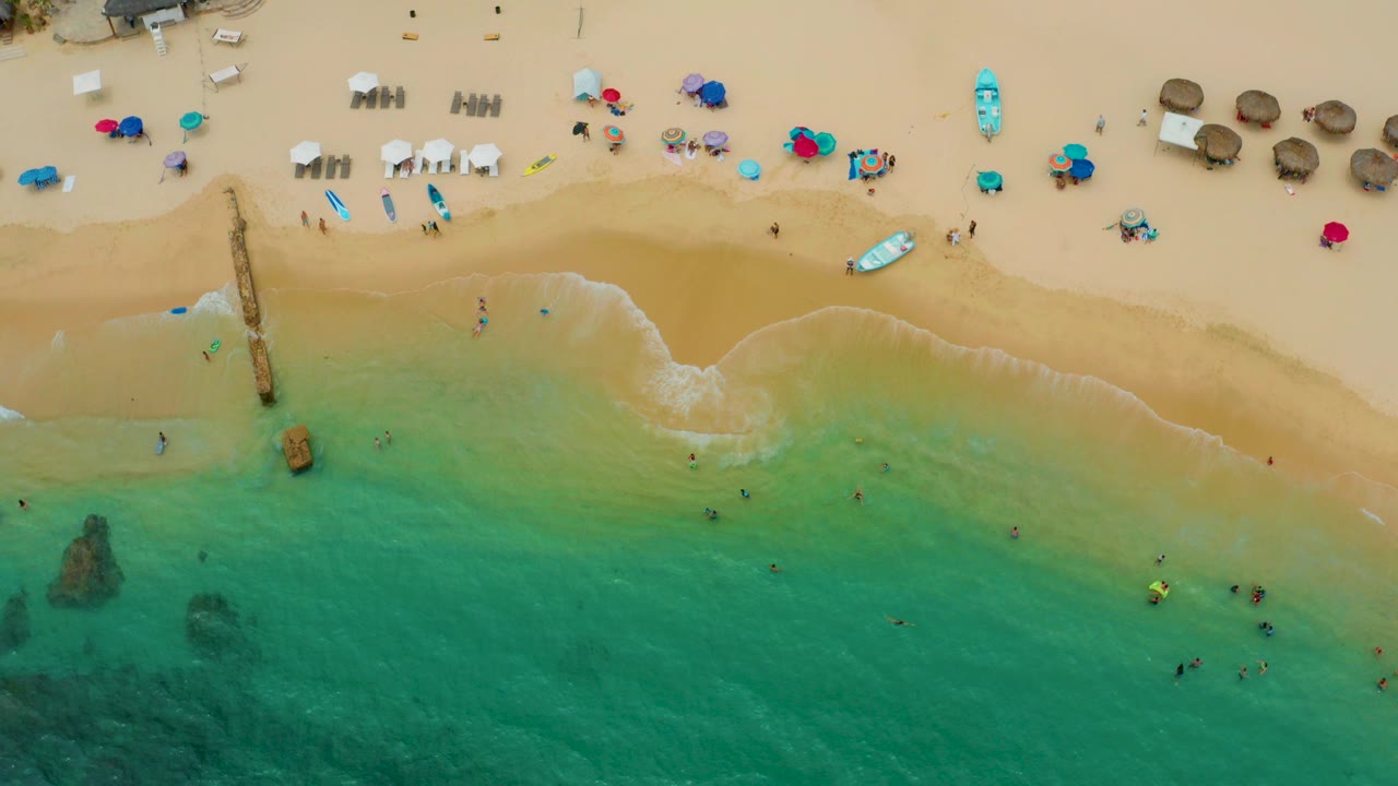 Top-down view of Palmilla Beach Cabo with green-blue shallows at the bottom, tan sand across the center, a narrow pier at left, and beach chairs and umbrellas along the upper shore.