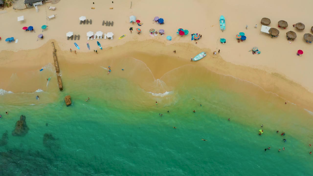 Palmilla Beach Cabo seen from above with the pier left of center, darker rocks under the water, pale surf on the sand, and clustered umbrellas and shade structures near the top of frame.
