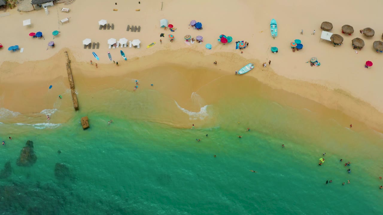 Overhead view of Palmilla Beach Cabo with a small foamy wave reaching the sand, a narrow pier on the left, and organized rows of beach chairs and umbrellas near the upper shoreline.