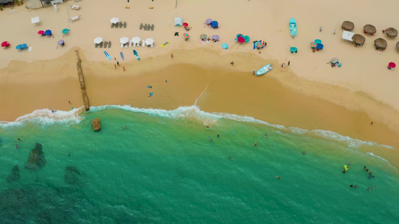 Overhead drone view of Palmilla Beach Cabo with tan sand, scattered umbrellas, people in the surf, and dark rock patches beneath turquoise water.