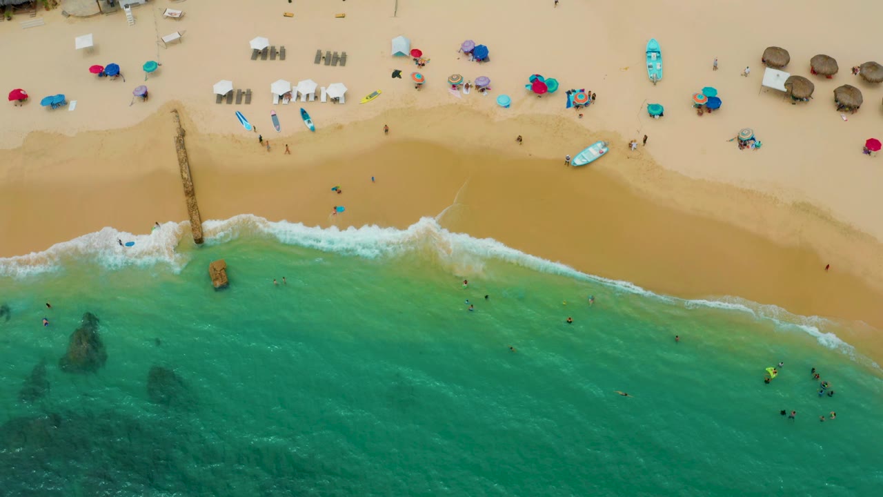Top-down Palmilla Beach Cabo shoreline with a diagonal band of surf, people in shallow water, umbrellas and chairs on sand, and darker shapes below the water surface.
