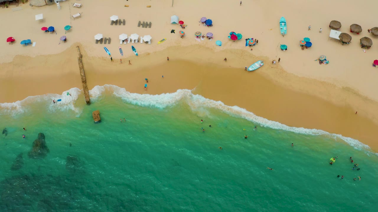 Drone view of sandy beach, surf, swimmers, and a narrow wooden structure.