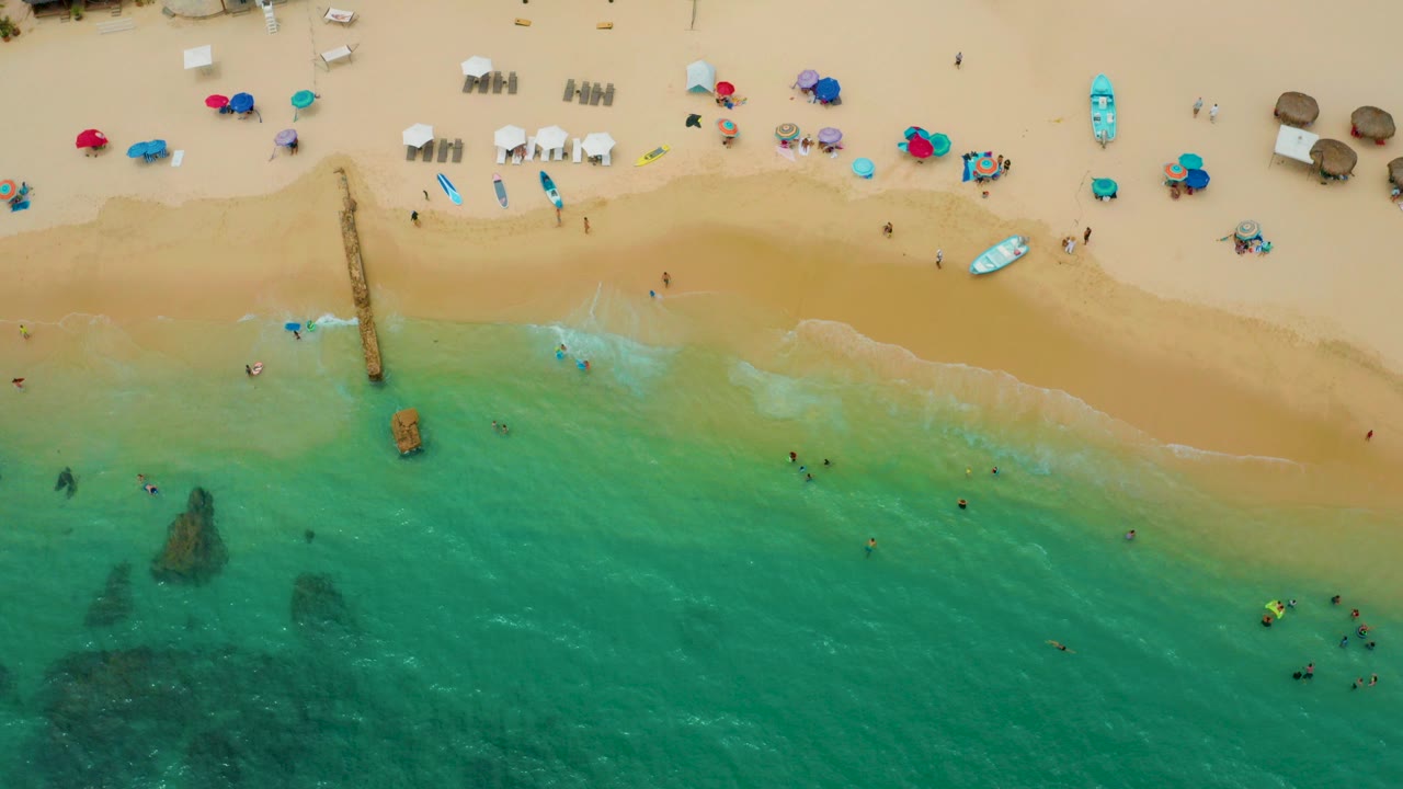 Overhead view of Palmilla Beach Cabo in Mexico with turquoise water at the lower half, tan sand above, umbrellas and chairs near the top, and small beachgoers in the surf.