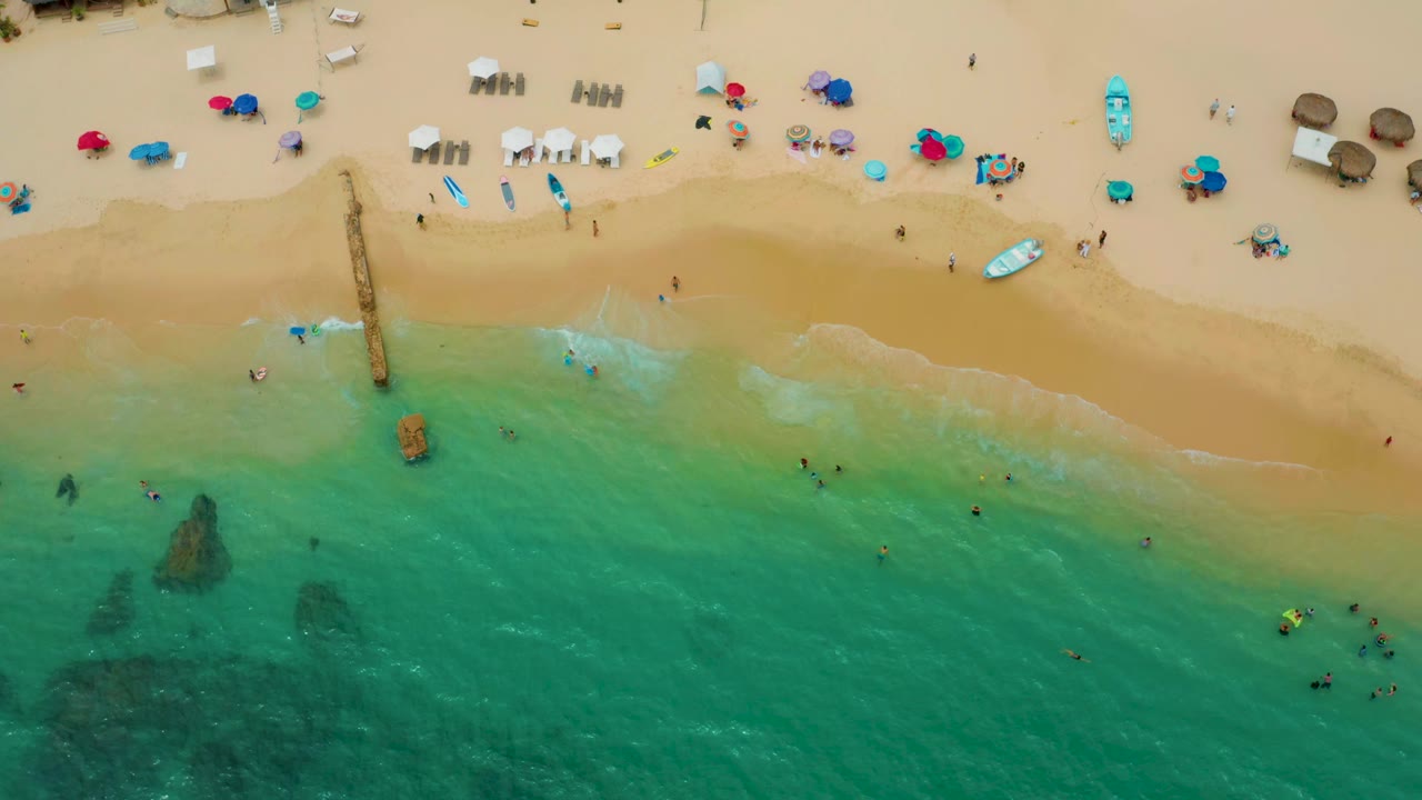 Aerial view over Palmilla Beach Cabo with swimmers scattered in shallow green-blue water, a sandy beach lined with umbrellas and chairs, and a narrow structure extending toward the surf.