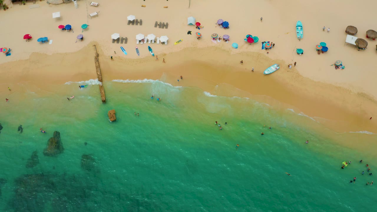 Drone-style overhead frame of Palmilla Beach Cabo showing the wave line along the sand, clusters of umbrellas and chairs above it, and small figures swimming or walking near the water.