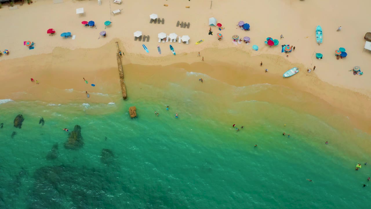 Drone view of beach umbrellas, surfboards, swimmers, and a narrow structure reaching into the surf.