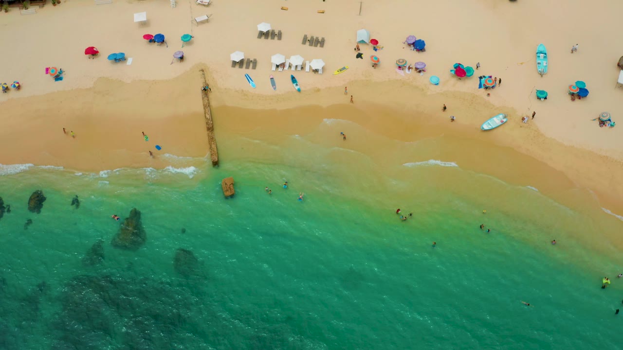 Palmilla Beach Cabo from above, showing pale surf breaking along the sand, several swimmers in the green-blue water, dark underwater rock patches, and clusters of umbrellas and chairs on the beach.