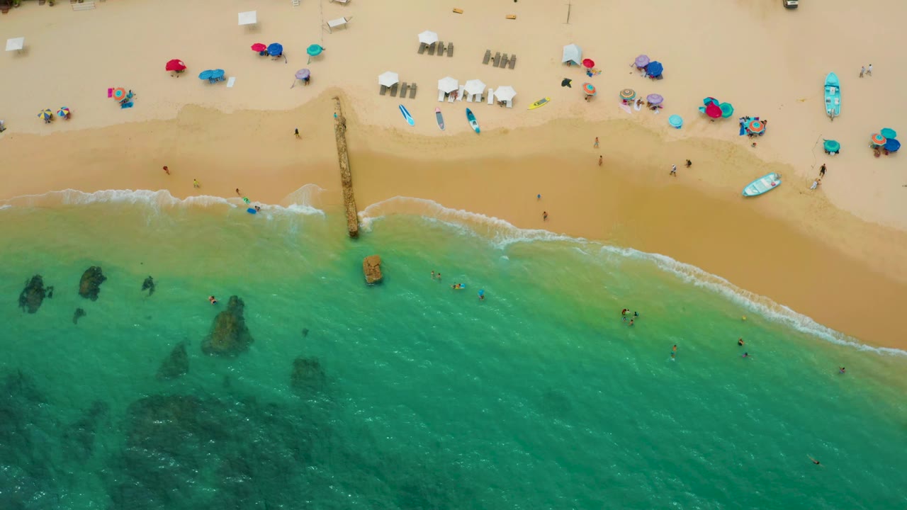 Palmilla Beach Cabo aerial frame showing green-blue shallows, a pale wave line, umbrellas and lounge chairs on the sand, and tiny swimmers visible below the pier area.