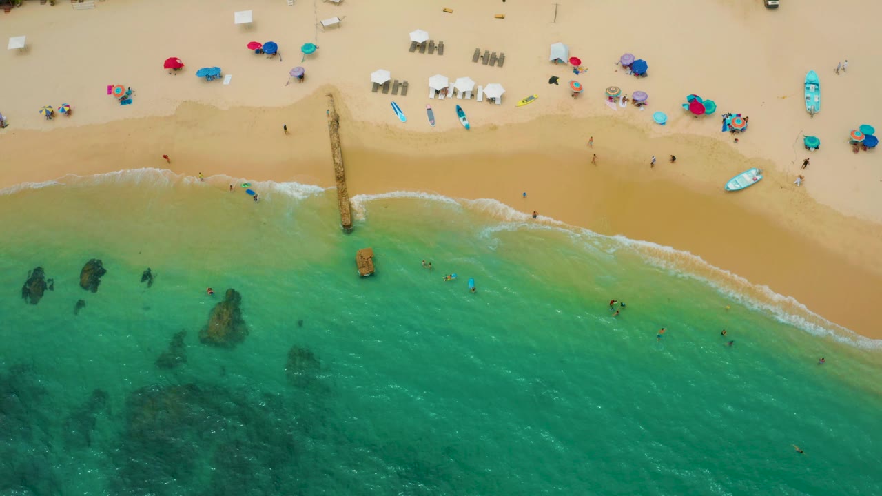 Top-down Palmilla Beach Cabo view with aqua water filling the lower frame, dark underwater rock shapes, a thin surf edge, scattered umbrellas, and tiny beachgoers on the sand.