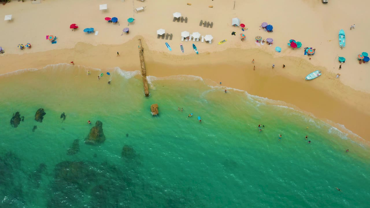 Drone-like overhead view at Palmilla Beach Cabo with the pier near center, turquoise shallows below, tan sand above, and small visible figures at the waterline.