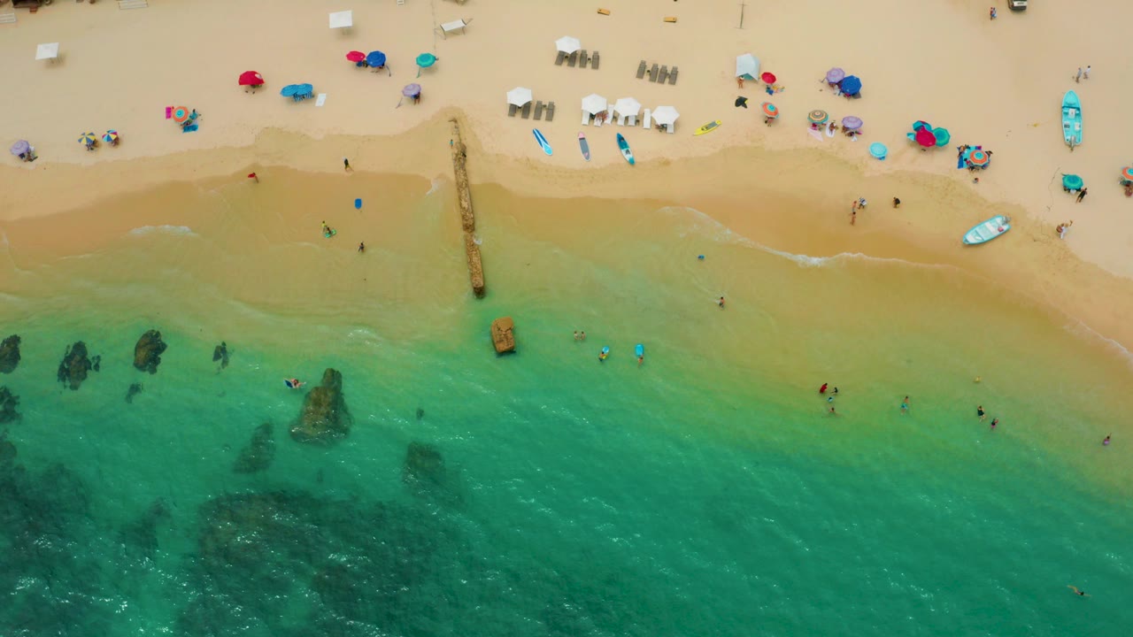 Aerial view of Palmilla Beach Cabo with turquoise water below, pale sand above, scattered beach umbrellas, boards or small boats, and tiny people swimming near shore.