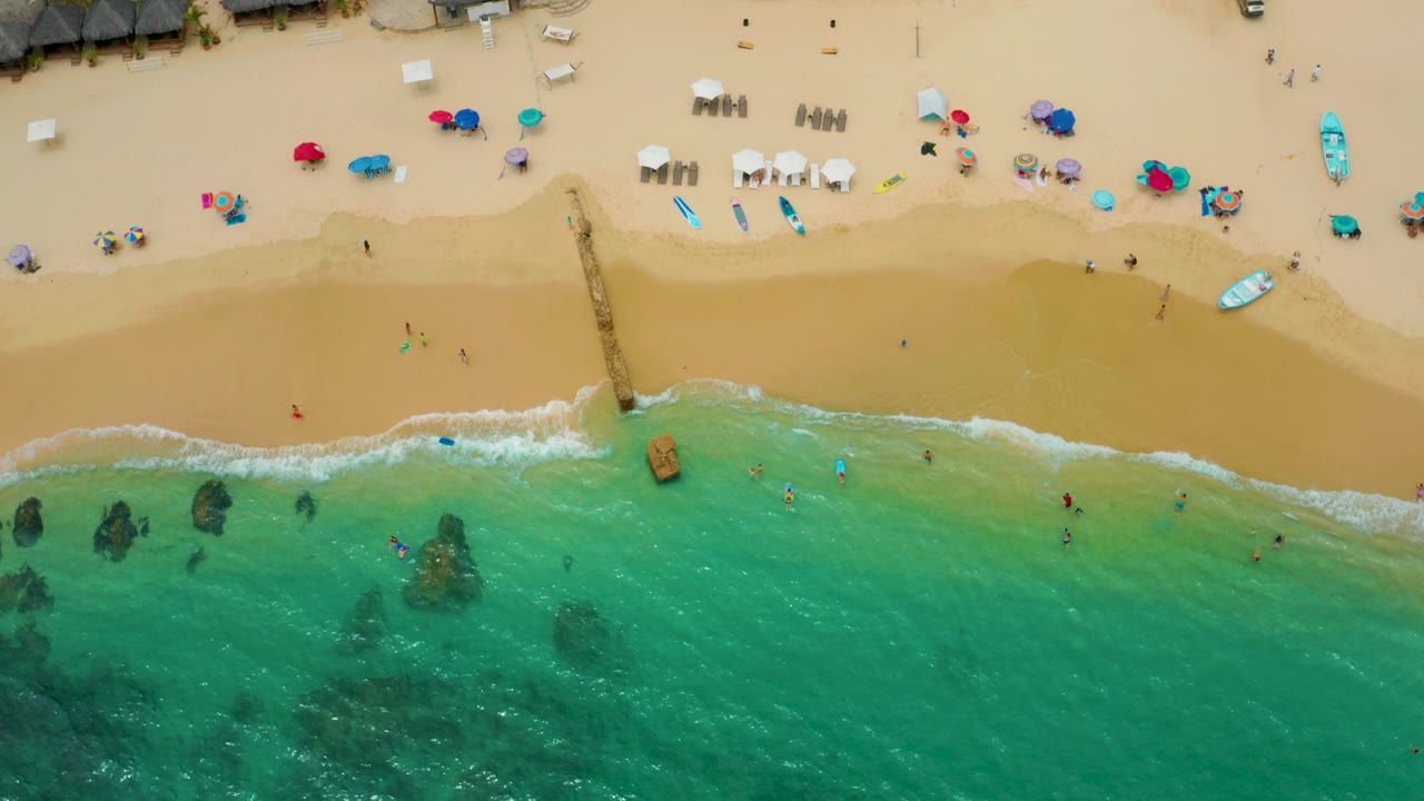 Overhead frame of Palmilla Beach Cabo, Mexico showing turquoise water below, golden sand above, a narrow pier near center, beach umbrellas and chairs, with tiny distant people not identifiable.