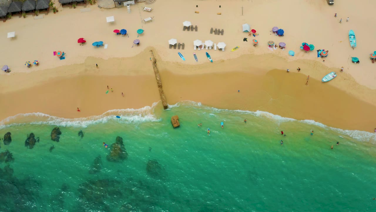 Top-down view at Palmilla Beach Cabo, Mexico with clear turquoise surf reaching the sand, a central pier, scattered umbrellas and lounge chairs, and tiny distant beachgoers or swimmers.