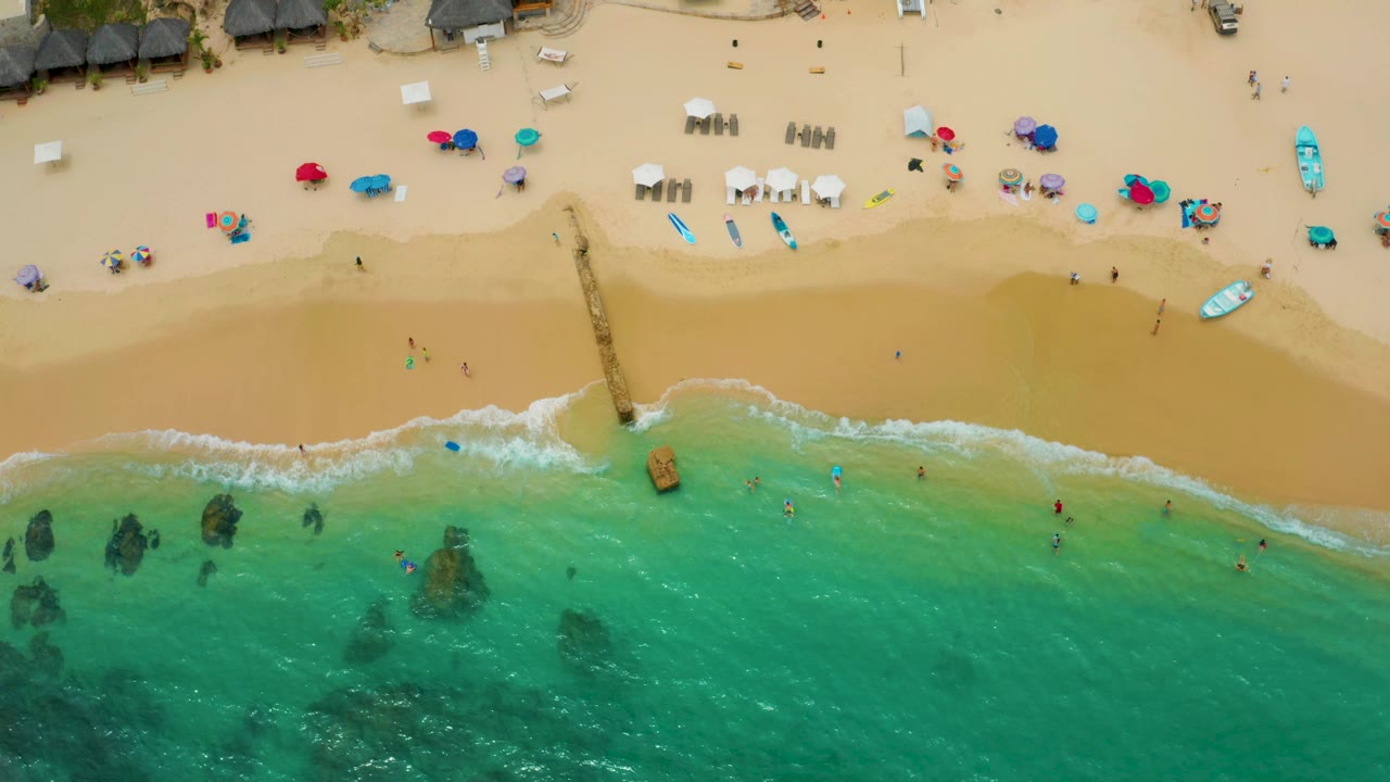 Aerial frame of Palmilla Beach Cabo, Mexico with shallow blue-green water, dark submerged rocks, a pier dividing the sand, and beach umbrellas and small boats along the upper beach.