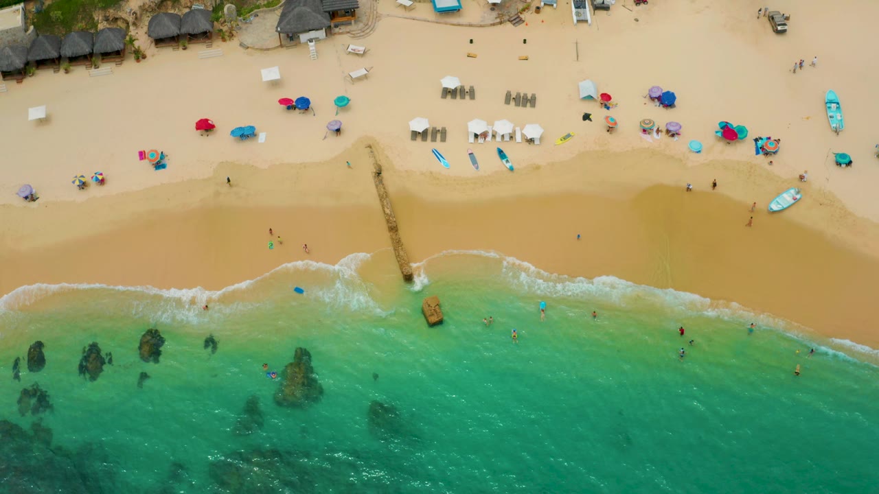 Overhead Palmilla Beach Cabo, Mexico frame with a broader band of pale wave wash, beach chairs and umbrellas above the waterline, small boats on the sand, and dark rock patches offshore.