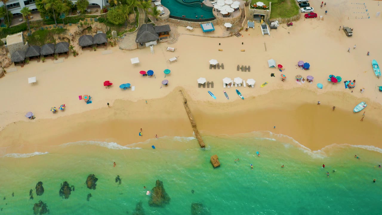 Overhead view of Palmilla Beach Cabo with tan sand, colorful umbrellas, palapa shade huts, resort buildings, swimmers, and turquoise water along the lower edge.