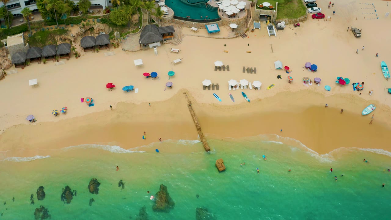 Aerial beach frame with a narrow groin, umbrellas, swimmers, and resort pool area.