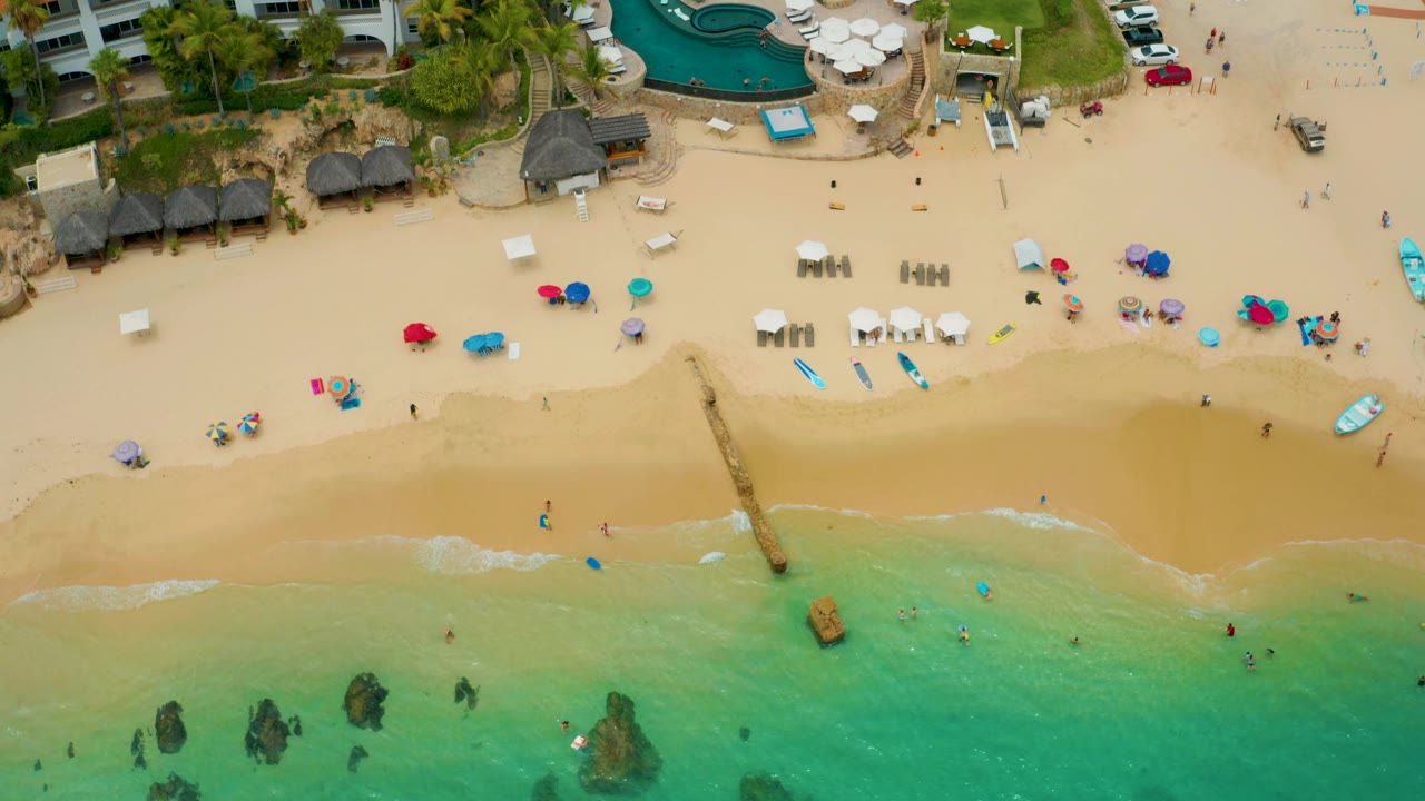 Aerial view over Palmilla Beach Cabo showing clustered umbrellas, white lounge chairs, palapa huts, people in the water, resort buildings, pool area, and parked cars at the upper right.