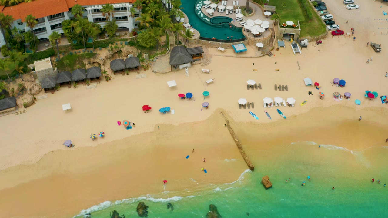 Overhead beach view with umbrellas, chairs, buildings, pool, and shallow surf.