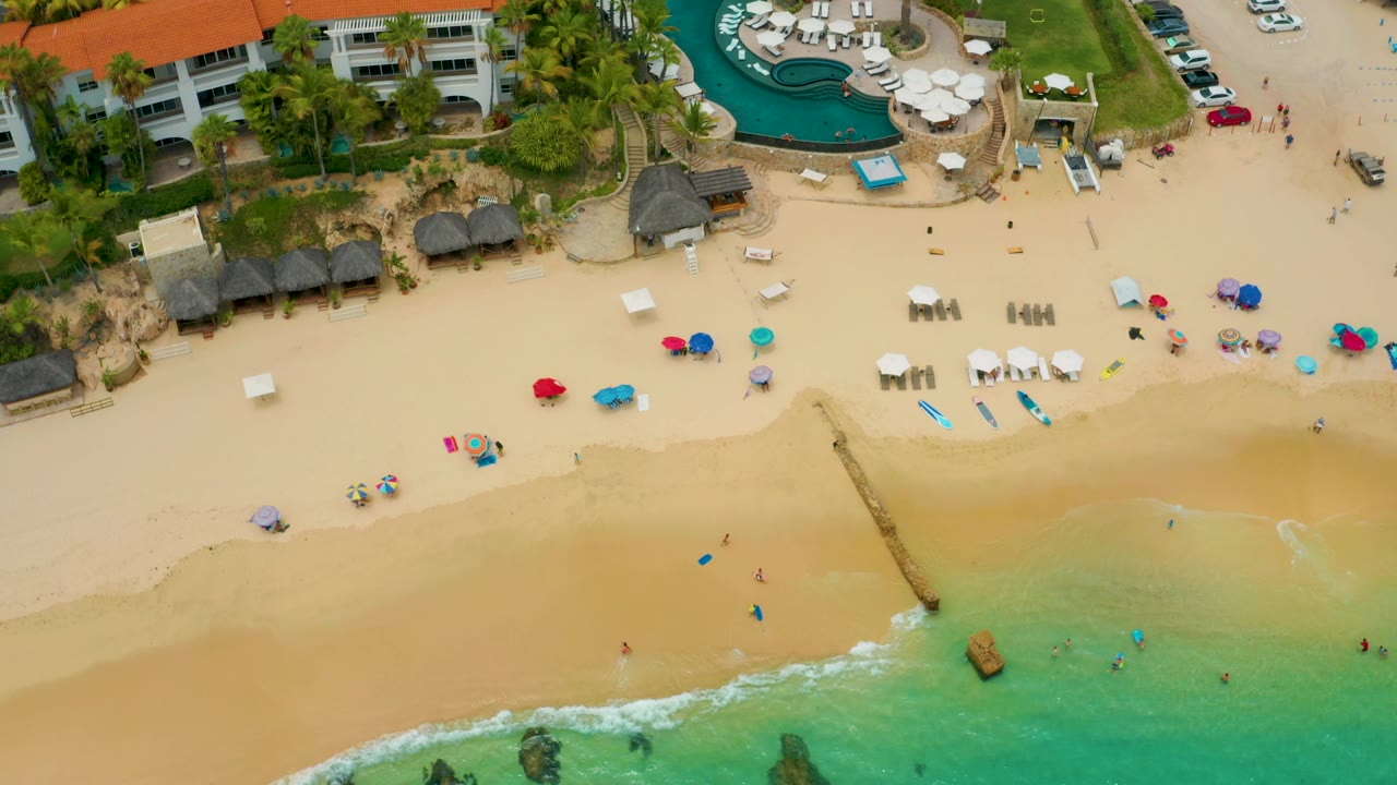 High aerial frame of Palmilla Beach Cabo, Mexico, showing coastal buildings, a curved pool, clustered umbrellas, open sand, light surf, and small distant figures near the waterline.