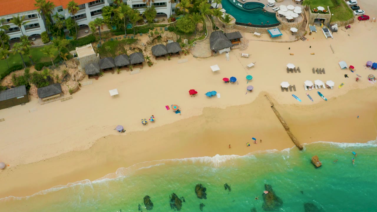 Aerial frame over Palmilla Beach Cabo, Mexico, with turquoise water at the bottom, pale sand through the center, thatched huts and palm-lined hotel grounds at the top, and tiny distant beachgoers.