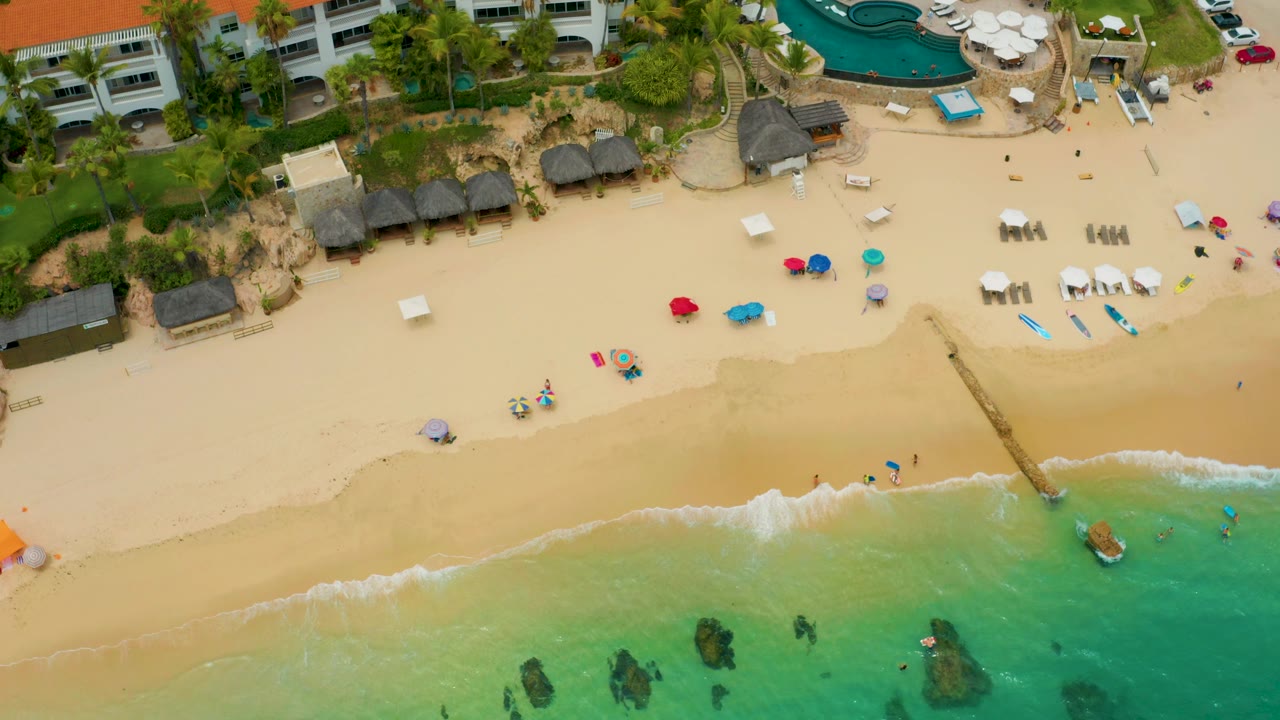 Palmilla Beach Cabo appears from above with shallow green-blue water and rocks in the lower frame, a thin foam line on the shore, umbrellas and loungers on sand, and a resort pool edge near the top.
