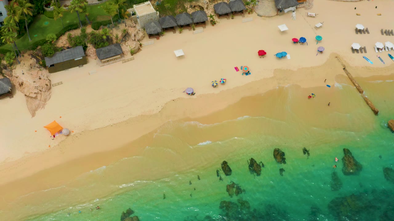 Aerial beach view with umbrellas, lounge areas, rocks in shallow water, and light surf.
