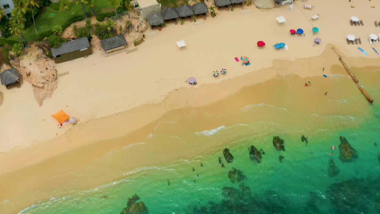 Top-down frame of Palmilla Beach Cabo, Mexico with the shoreline running diagonally, clear green water below, umbrellas on sand above, and no clearly identifiable people.