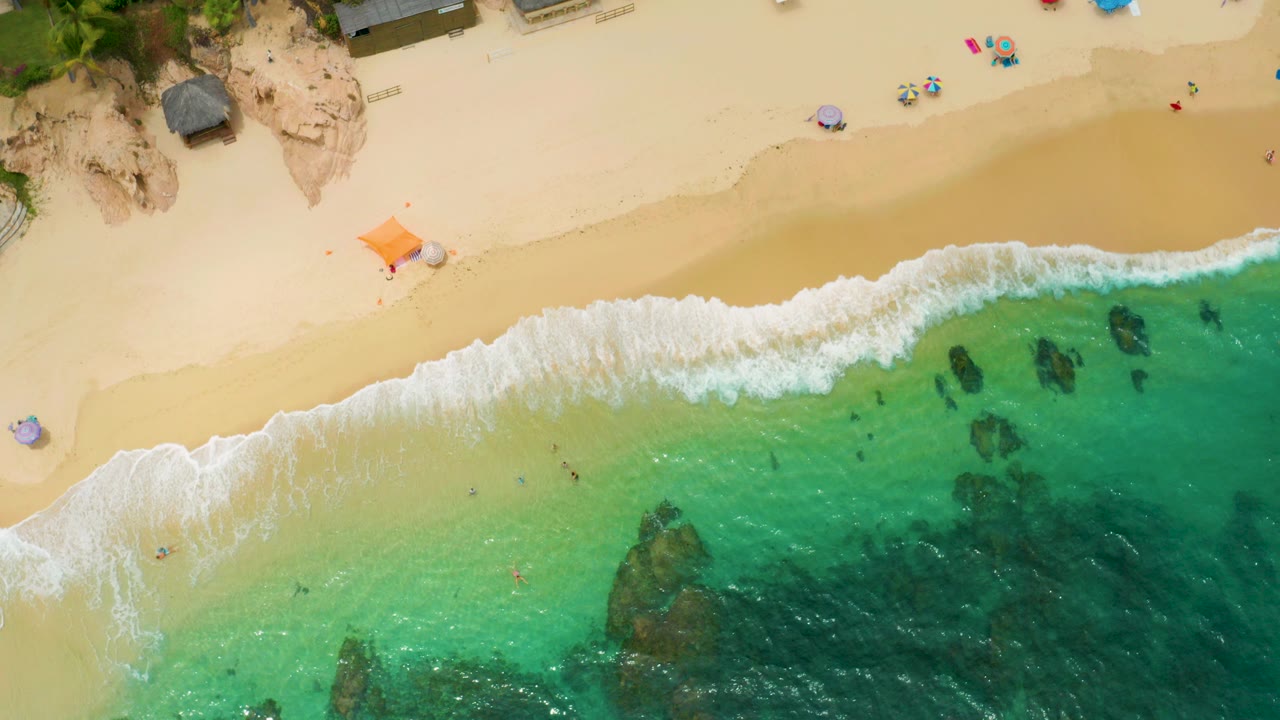 Overhead view of Palmilla Beach Cabo with pale sand, a white surf line, turquoise shallows, dark underwater rocks, beach umbrellas near the top edge, and no visible people.