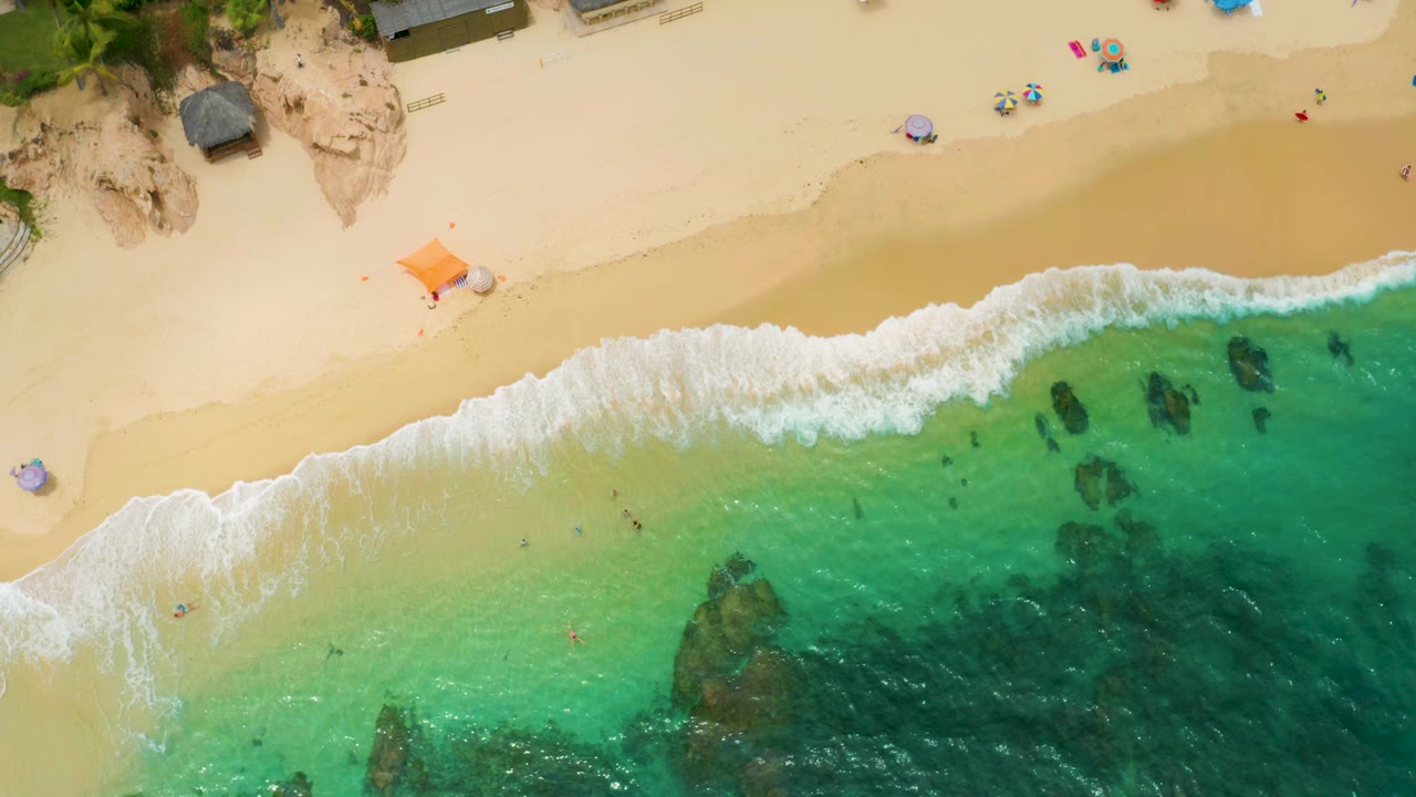 Palmilla Beach Cabo from above, with the surf line slightly farther across the frame, shallow green-blue water below it, dark rock patches, and no visible people.