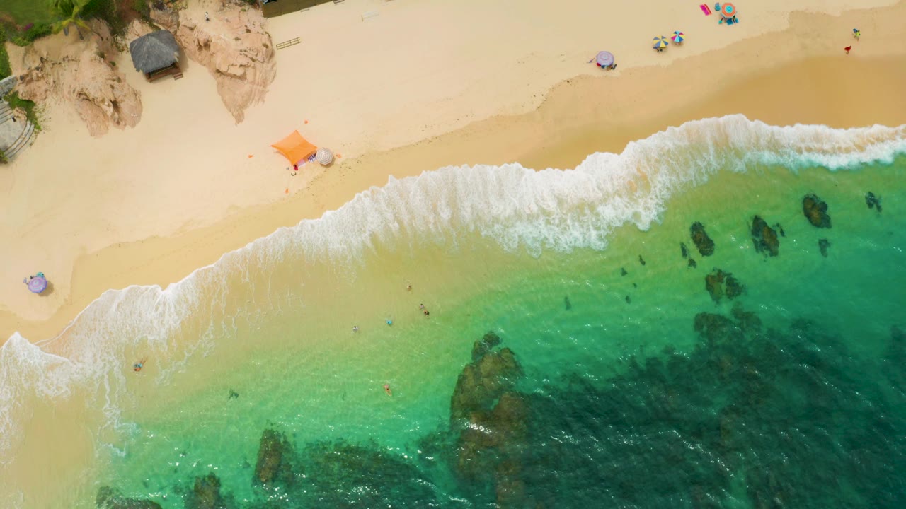 Overhead frame of Palmilla Beach Cabo showing a stronger white wave band on the sand, transparent turquoise water, scattered submerged rocks, small beach items, and no visible people.