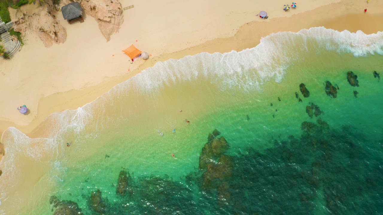 Top-down beach view with surf, sand, and dark rocks under water.